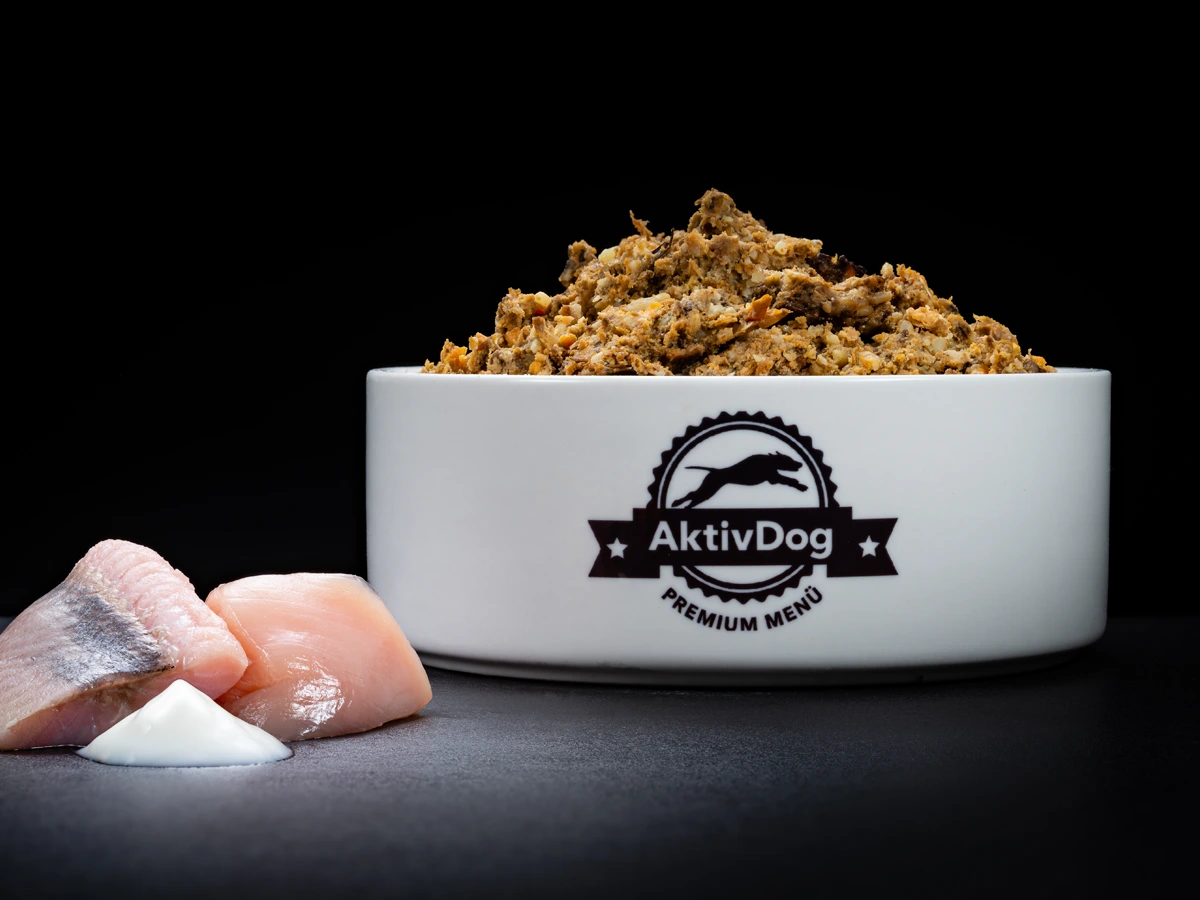 Großaufnahme von AktivDog Welpenfutter mit links daneben liegendem rohen Lachs, Geflügelfleisch und Joghurt